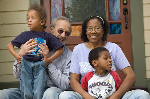 Grandparents smiling and holding their two grandchildren in front of their house.