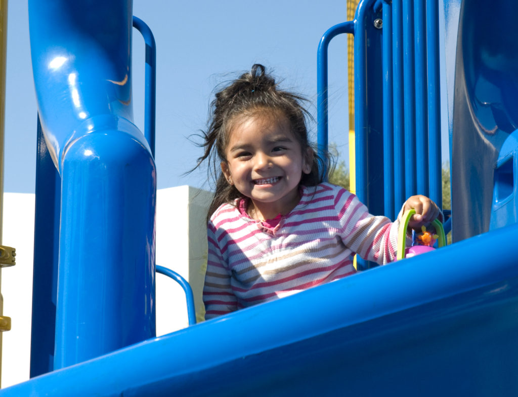 A child smiles on a blue slide while holding her toy.