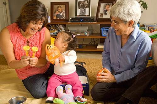 A mother plays with her child, who smiles widely while holding a slinky. A TLG Therapist sits nearby, also smiling.