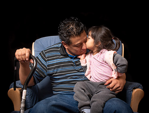 A young girl kisses the cheek of her father. Her father's eyes are closed and he holds a walking cane.