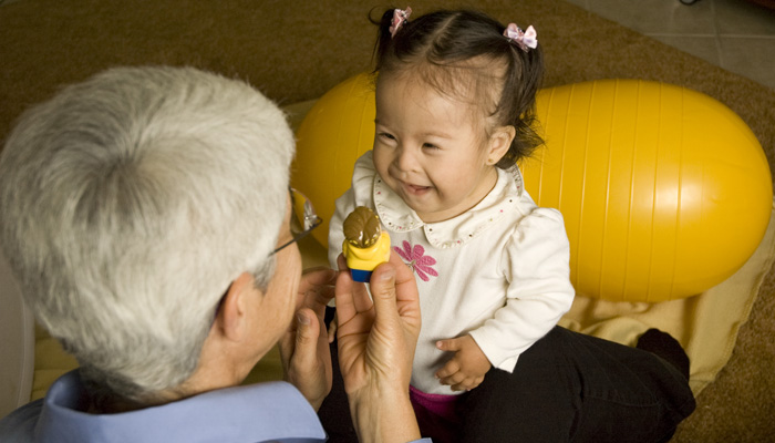A TLG therapist plays with a young child, showing her a figure from a block playset.